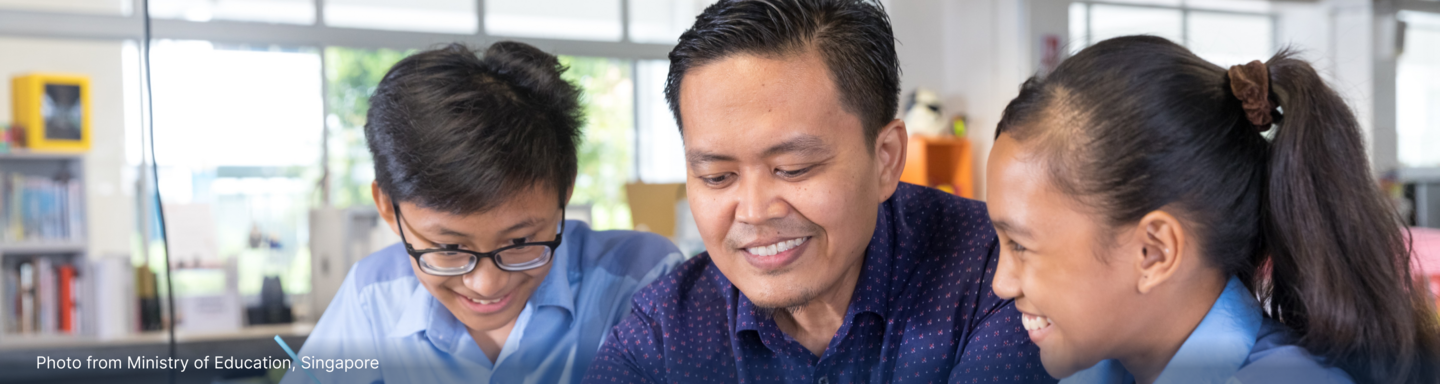 A smiling adult man sits between two students in a classroom, all looking down together at something.